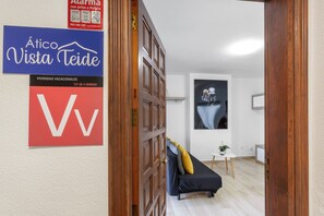 A hotel interior with a wooden door, a blue sign with white text, a red sign with a white letter, a black sofa with cushions, a small table with a plant, and a wall-mounted picture. - Vacation Apartment 'Precioso Atico Vista Teide' with Mountain View, Wi-Fi and Air Conditioning (Arona)