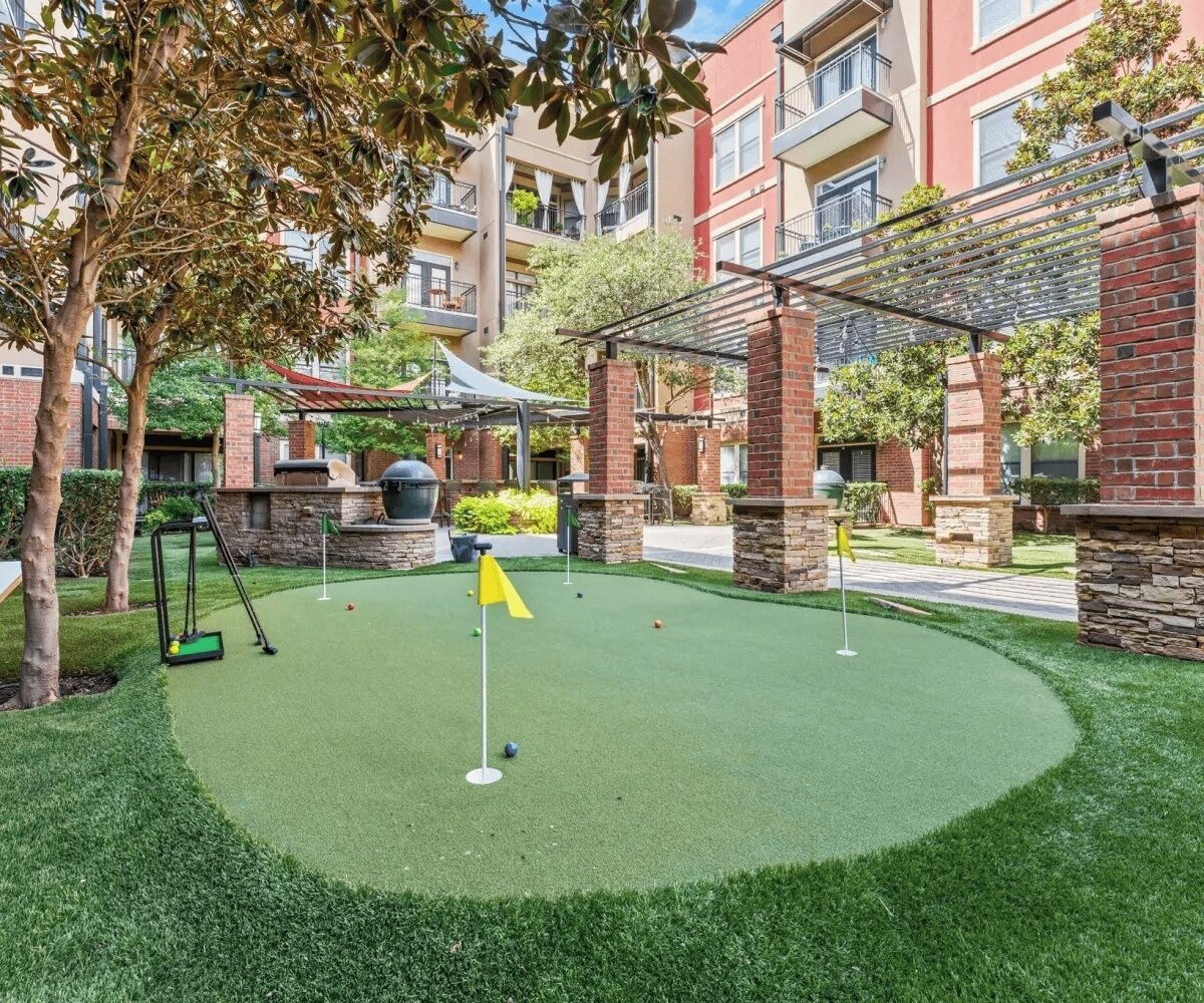 A miniature golf course with a green artificial turf, surrounded by brick buildings and trees.