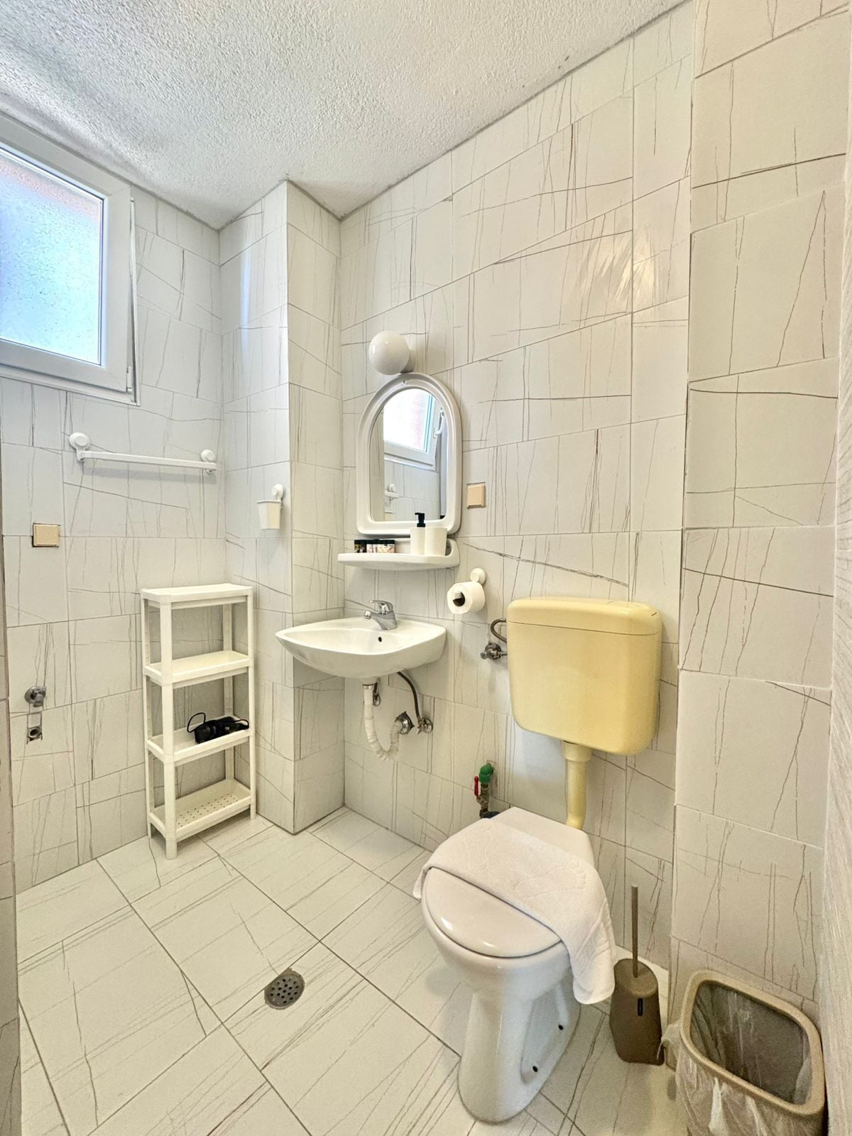 A small bathroom with a toilet, sink, and mirror. There is a shelf with toiletries and a window above the sink.