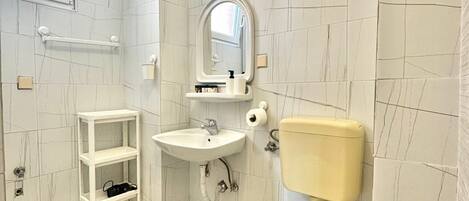 A small bathroom with a toilet, sink, and mirror. There is a shelf with toiletries and a window above the sink.