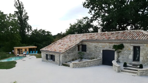 A stone building with a tiled roof, a swimming pool, and a small wooden structure with blue chairs.
