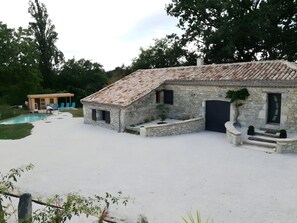 Un edificio de piedra con techo de tejas, una piscina y una pequeña estructura de madera con sillas azules.