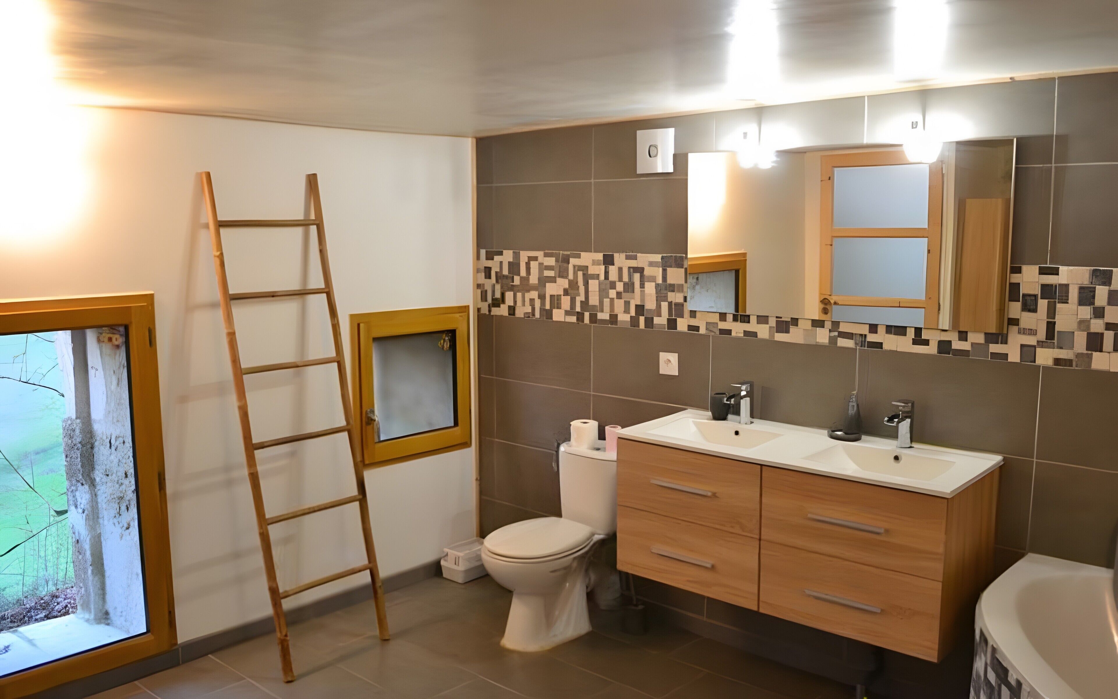A bathroom with a toilet, sink, vanity, and a bathtub. There is a window with a view of trees and a ladder leaning against the wall.