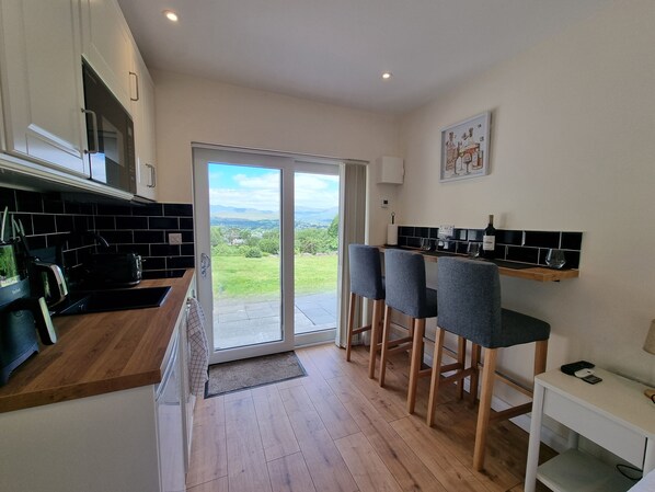 A modern kitchen with a sliding glass door leading to a balcony, wooden flooring, and bar stools. - Misty Bay (Dawros)