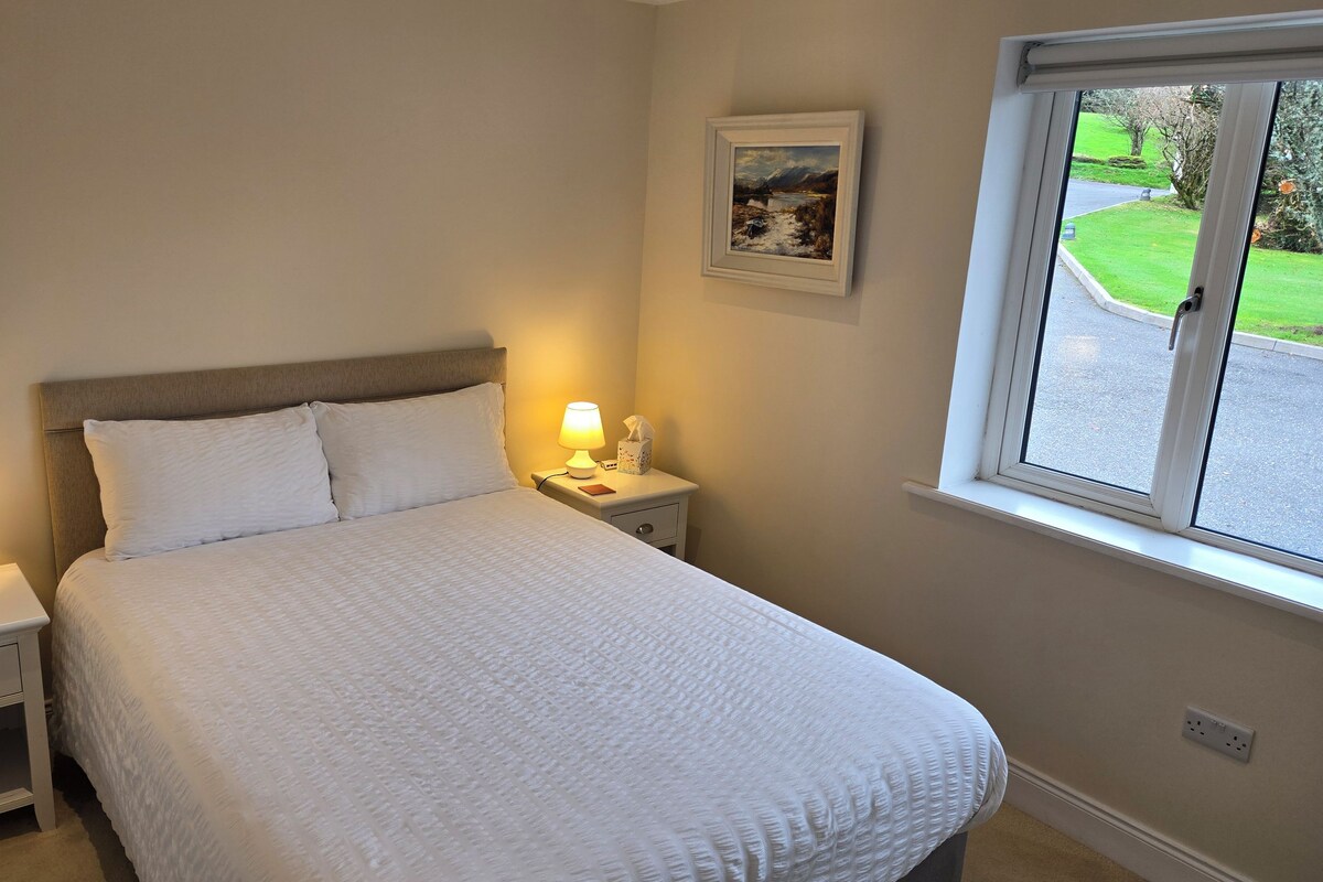 A neatly made bed with white linens, two bedside lamps, a bedside table with a tissue box, and a framed landscape painting on the wall.