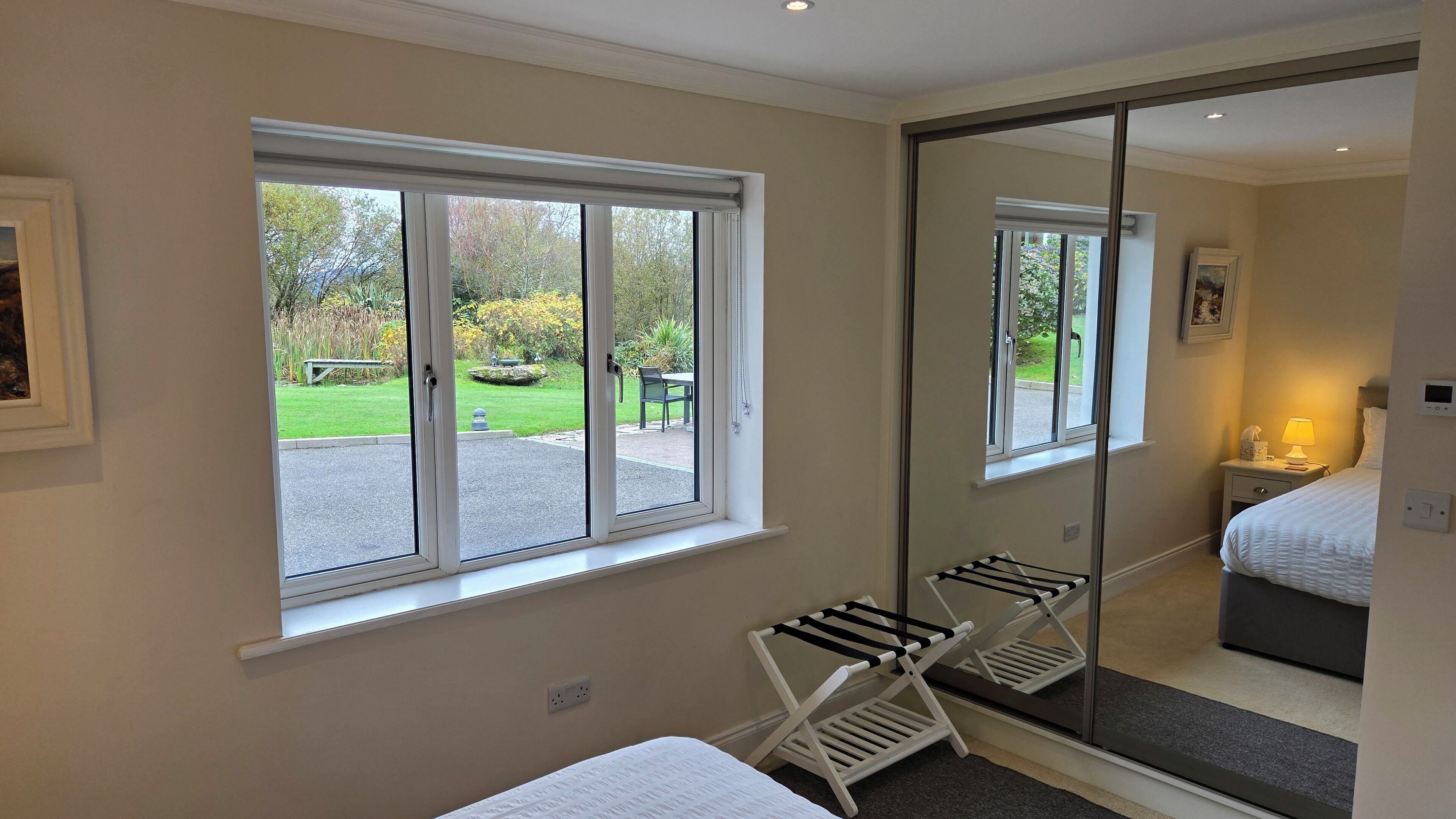A bedroom with a large mirror, a bed, a bedside table with a lamp, and a window with a view of a garden.