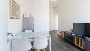 Un salon moderne avec une table en verre, des chaises blanches, un téléviseur à écran plat, un meuble en bois foncé et un tableau encadré au mur.