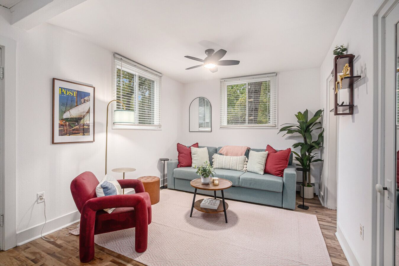 A living room with a red chair, a blue sofa, and a ceiling fan. There are two windows with blinds, a floor lamp, and a plant.