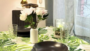 Ein Esstisch mit einer grün gemusterten Tischdecke, einer Vase mit weißen Blumen, einem schwarzen Teller und zwei Gläsern.