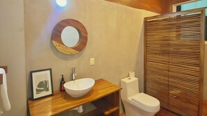 Een moderne badkamer met een houten wastafelmeubel, een ronde spiegel, een witte wasbak, een toilet en een houten privacywand.