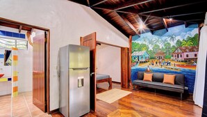 Um quarto com piso de madeira, sofá, geladeira e um mural com tema tropical.