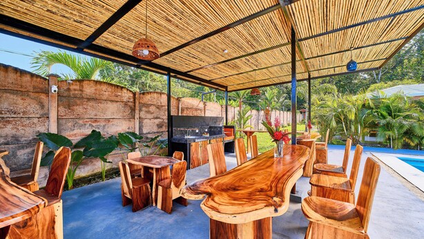Uma área de refeições ao ar livre com teto de palha, móveis de madeira e piscina ao fundo.