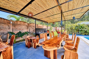 An outdoor dining area with a thatched roof, wooden furniture, and a pool in the background. - Villas Ande (Puerto Viejo de Talamanca)