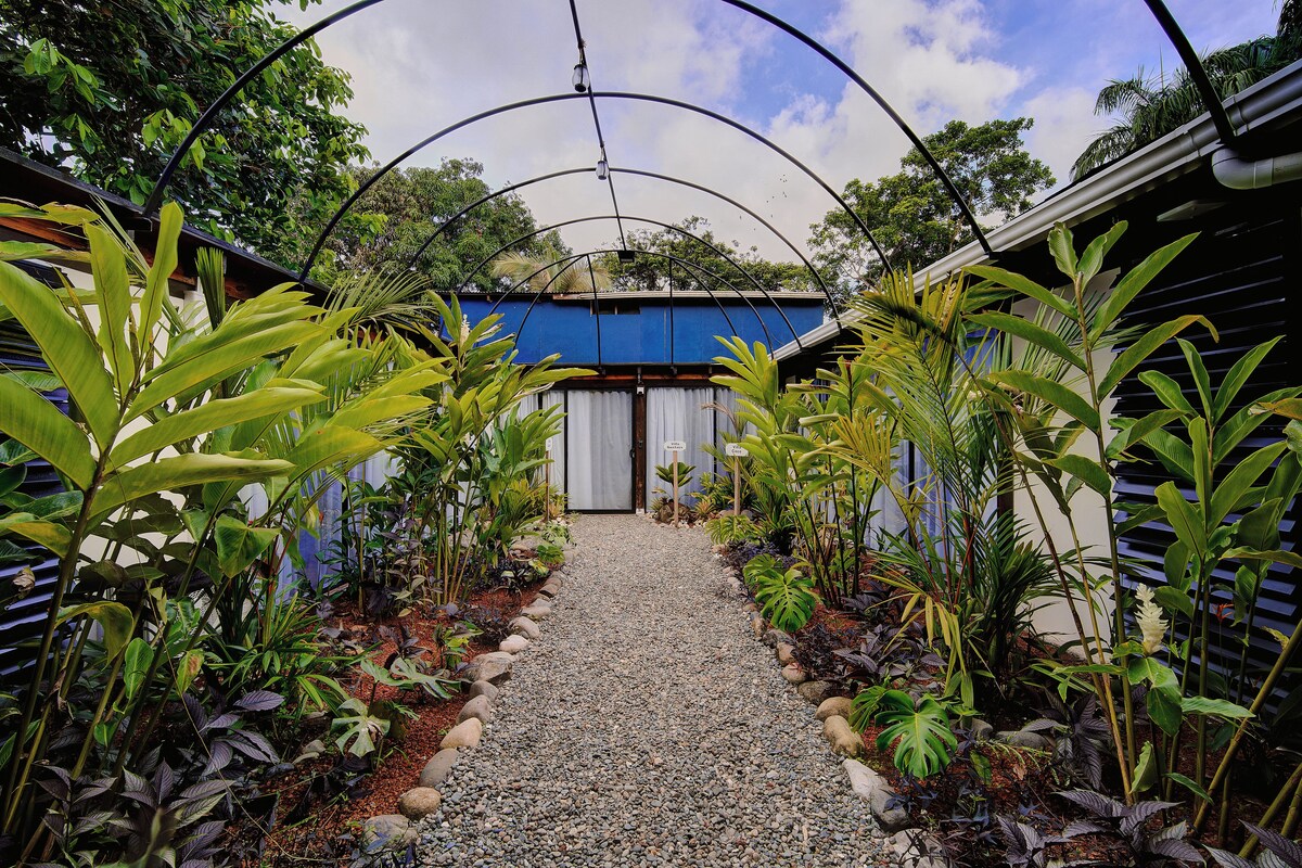 Un chemin menant à un bâtiment surmonté d’un toit bleu, entouré de plantes tropicales et bordé d’un sentier de gravier.