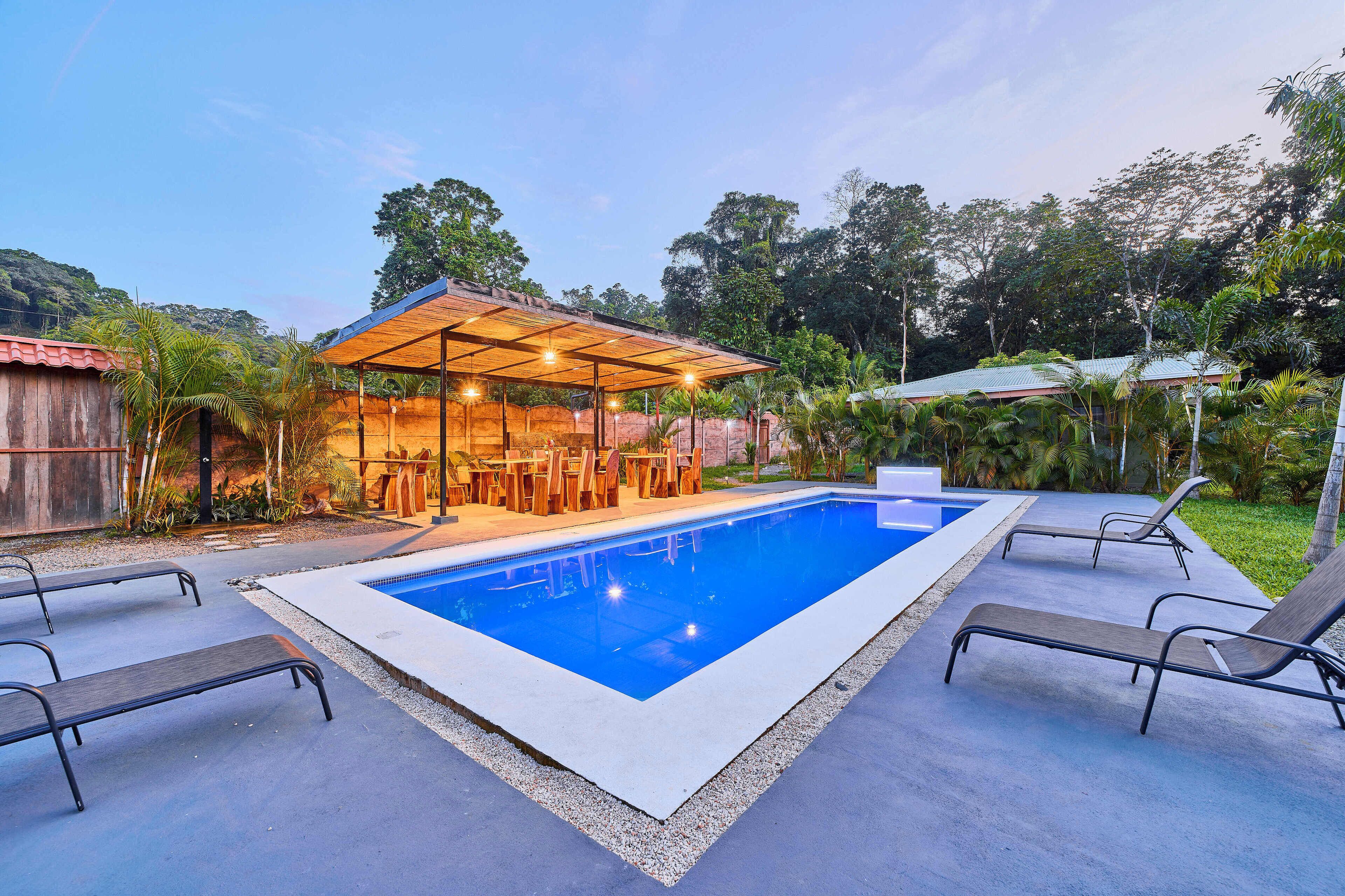 A swimming pool with lounge chairs, a covered seating area, and lush greenery in the background.