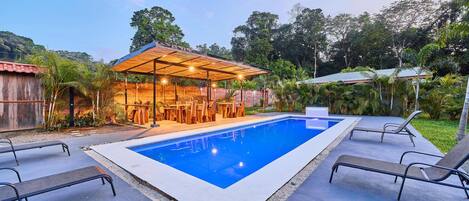 A swimming pool with lounge chairs, a covered seating area, and lush greenery in the background.