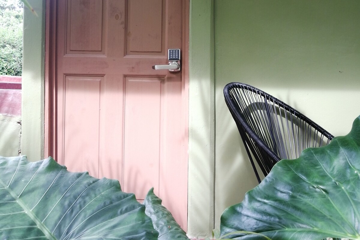 Une porte en bois dotée d’un clavier pour le verrouillage, une plaque fixée au mur portant l’inscription « Villa Tucan », et une chaise en métal noir.