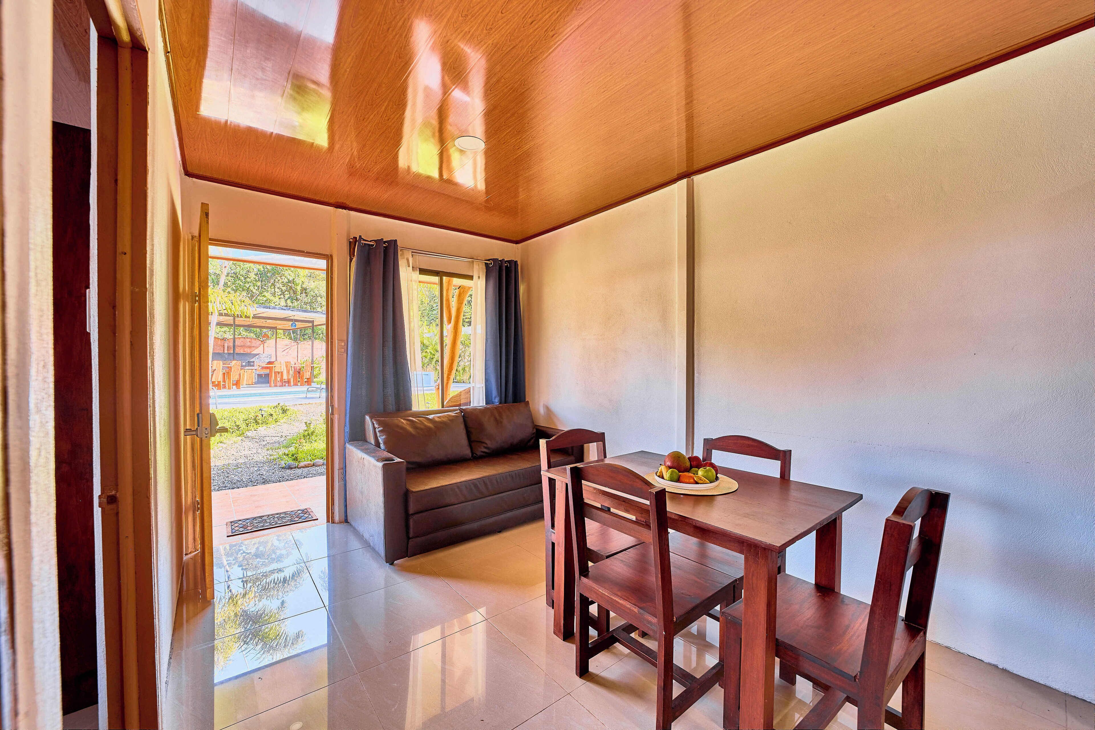 A modern room with a wooden ceiling, a dining table with chairs, a sofa, and a view of the outdoors.