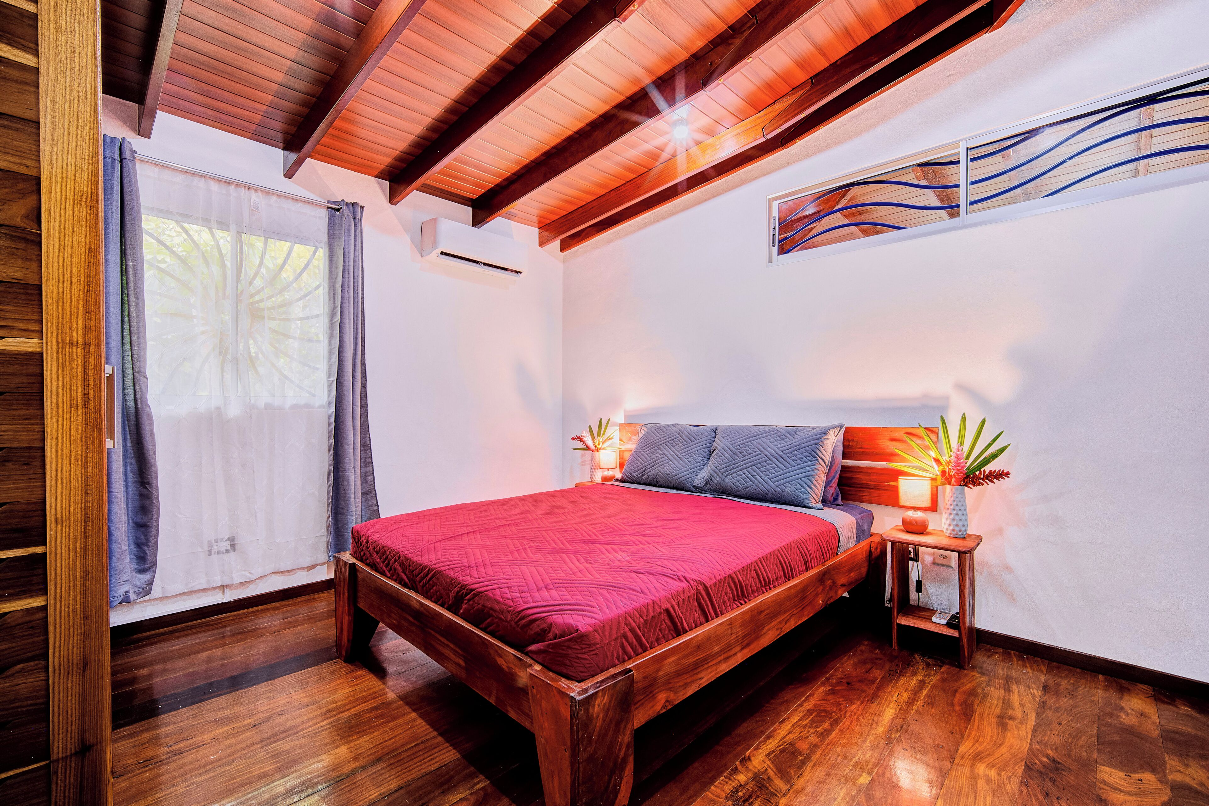 A bedroom with a wooden ceiling, a bed with a red bedspread, a nightstand with a lamp, and a window with curtains.