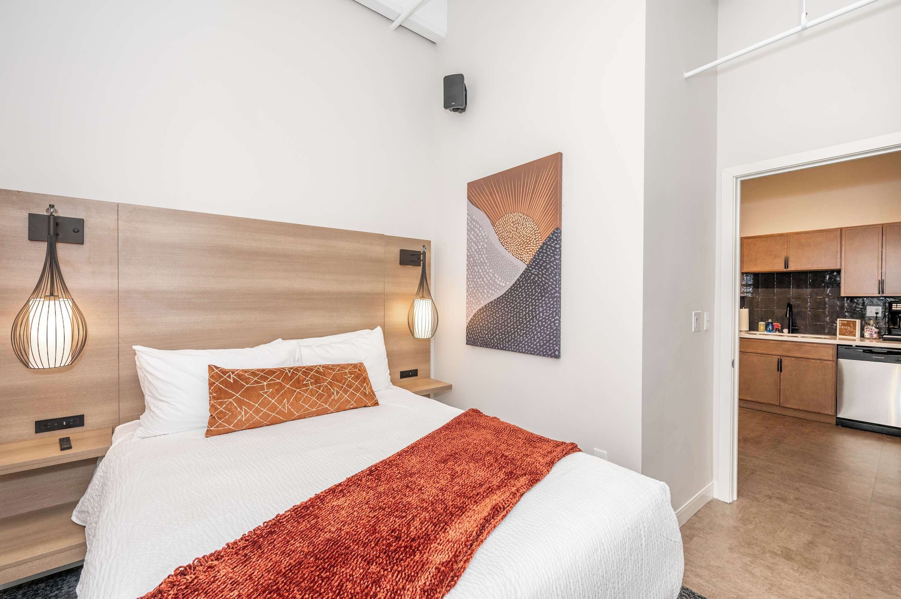 Una habitación de hotel moderna con cabecera de madera, una cama grande con ropa de cama blanca y naranja, una manta roja y una lámpara de pared. Se ve una cocina a través de una puerta abierta.