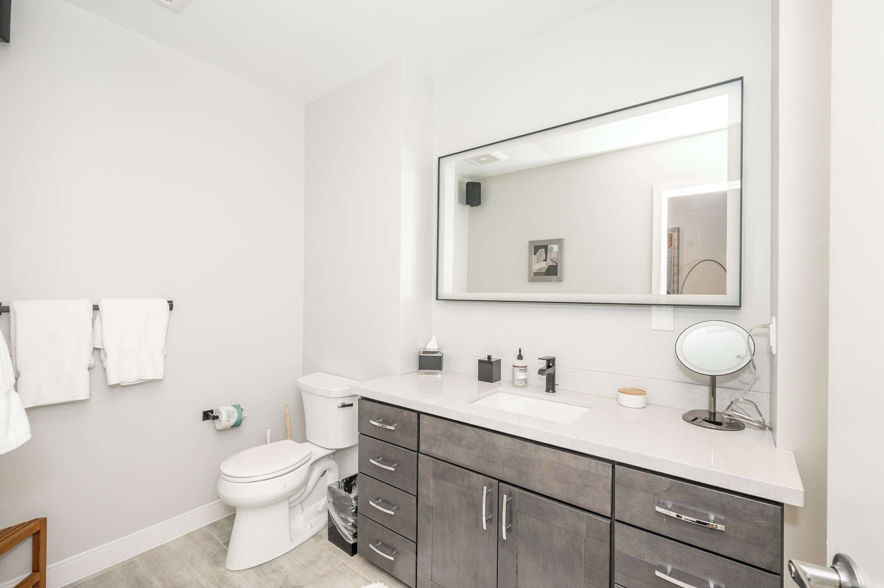 A modern bathroom with a white toilet, grey cabinets, a large mirror, and a white sink.