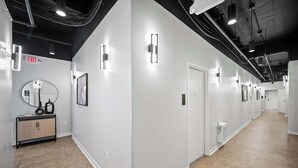 Ein Gang mit weißen Wänden, Holzboden und schwarzer Decke mit freiliegenden Rohren. Wandleuchten und ein Ausgangsschild sind vorhanden.
