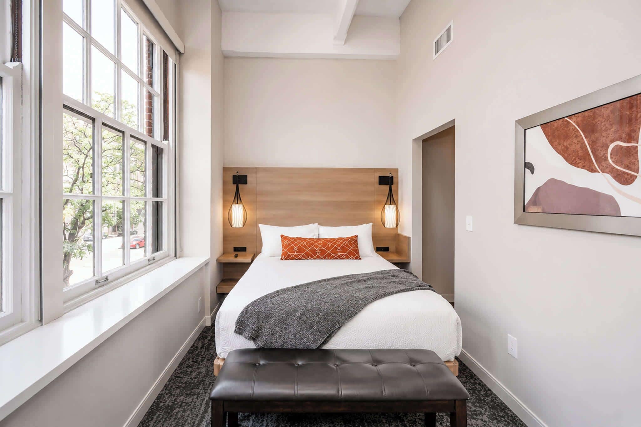 A hotel room with a bed, bedside lamps, a bench, and a window.