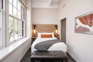 Una habitación de hotel con una cama, lámparas de noche, un banco y una ventana.