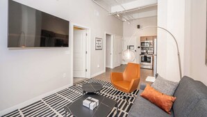 Ein modernes Wohnzimmer mit einer Couch, einem Couchtisch und einem orangefarbenen Sessel. Ein großer Flachbildfernseher ist an der Wand befestigt.