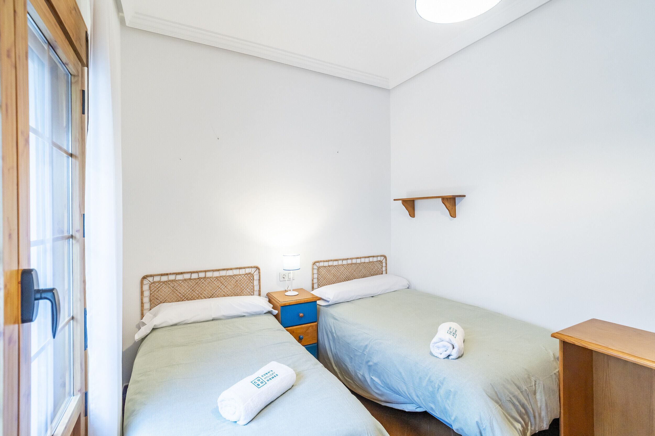 Una habitación con dos camas individuales, una mesita de noche de madera con una lámpara, un estante pequeño y una ventana con persianas.