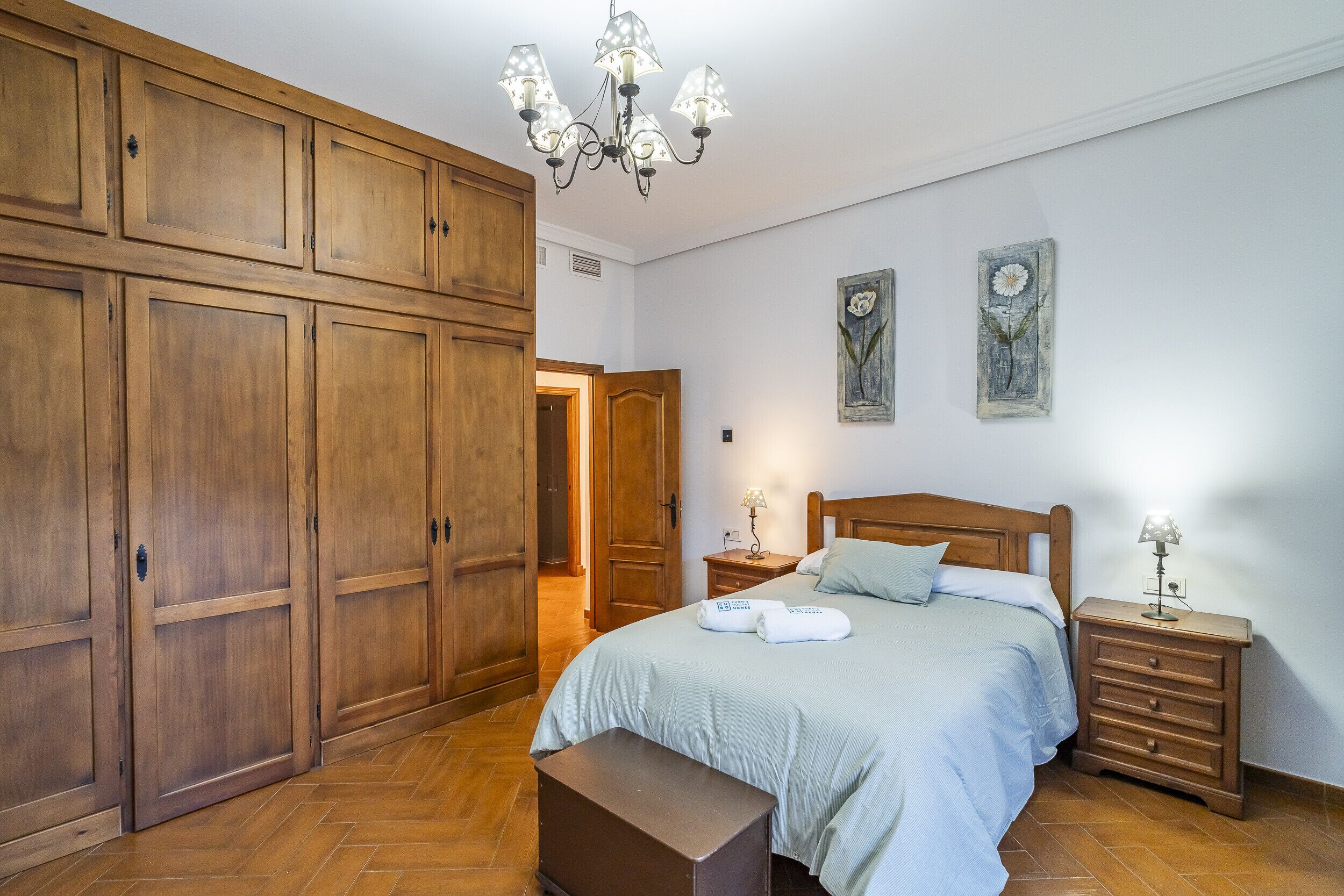 Un dormitorio con un amplio armario de madera, una cama con dos almohadas, una mesita de noche con lámpara y un banco.