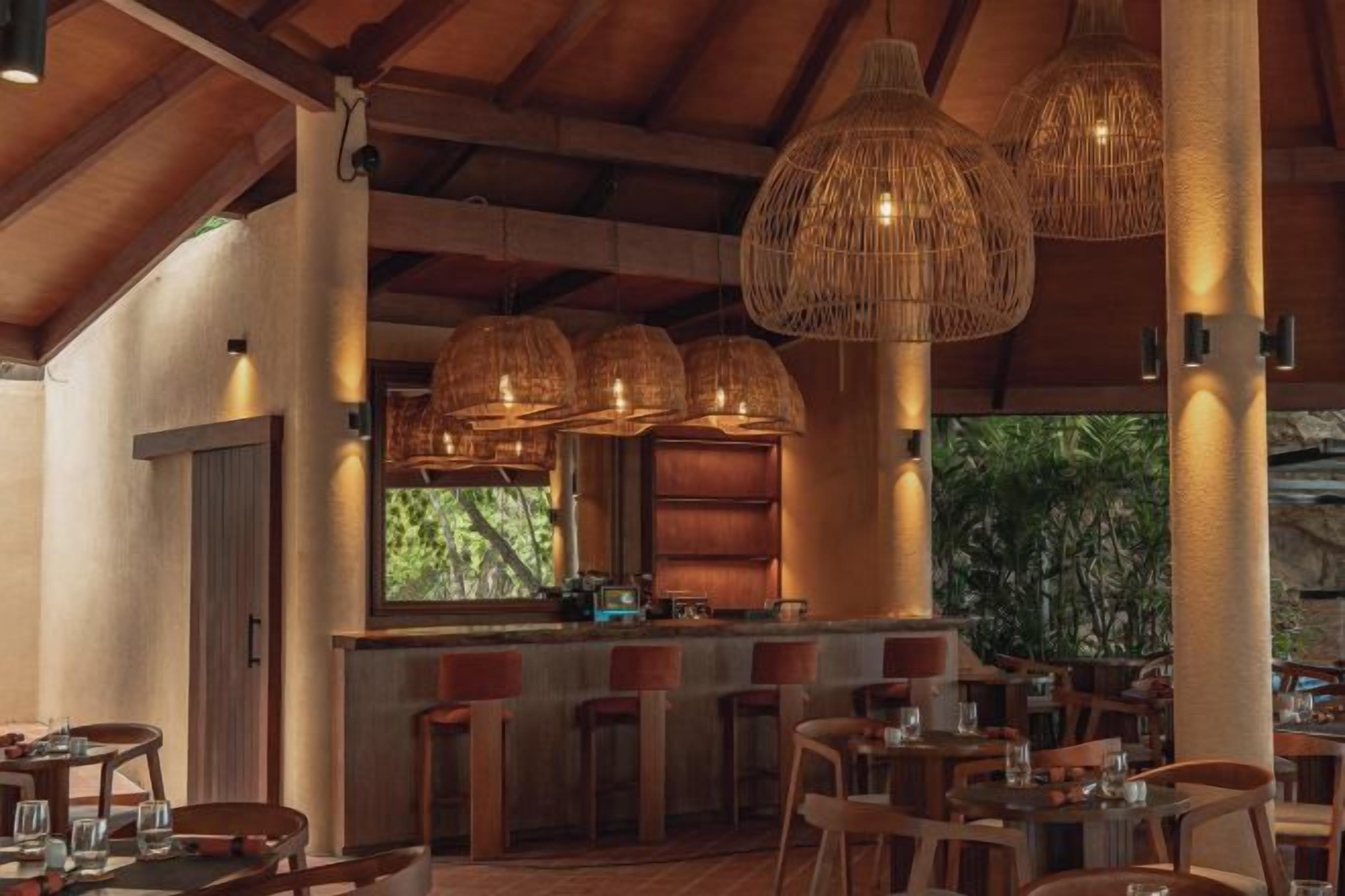 Un restaurante con un interior rústico, que cuenta con vigas de madera, lámparas colgantes de mimbre y una zona de bar con taburetes.
