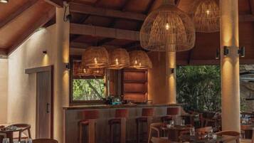 Un restaurante con un interior rústico, que cuenta con vigas de madera, lámparas colgantes de mimbre y una zona de bar con taburetes.