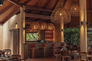 Een restaurant met een rustiek interieur, met houten balken, hangende wilgentaklampen en een bar met krukken.