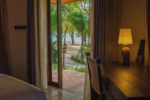 Een kamer met een bed, een bureau, een lamp en uitzicht op een tropisch buitengebied met een pad en palmbomen.