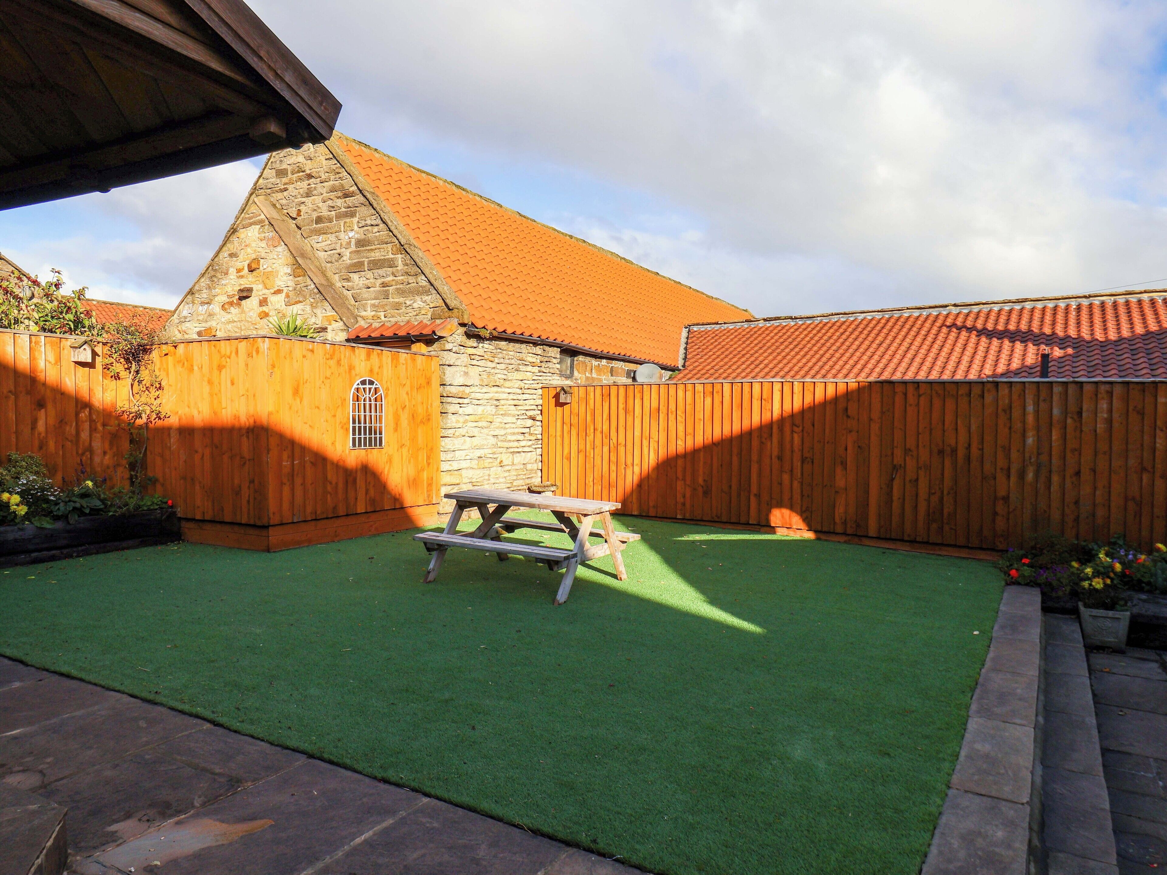 Un patio trasero con césped artificial verde, una mesa de picnic de madera y un edificio de piedra con tejado de tejas rojas.