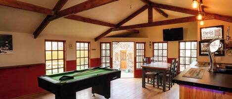 A pool table in a room with wooden beams, a bar area with a tap, and large windows.