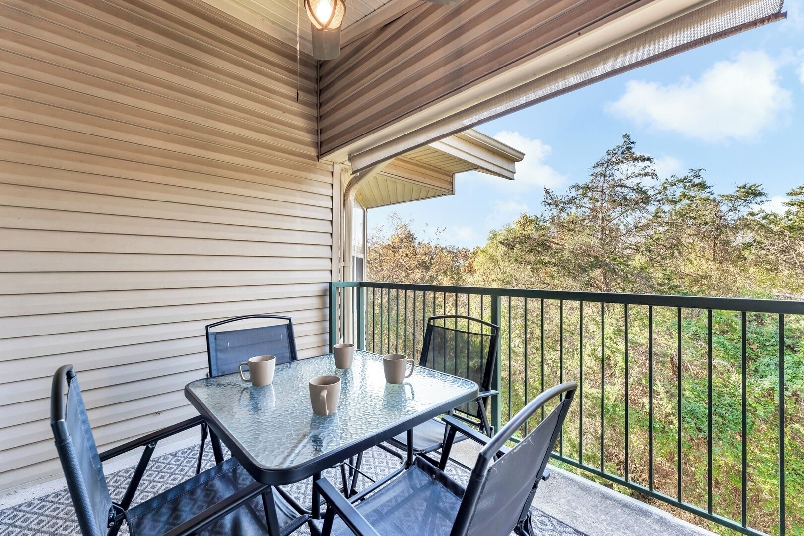 A covered patio with a table and chairs, a railing, and a view of trees and sky.
