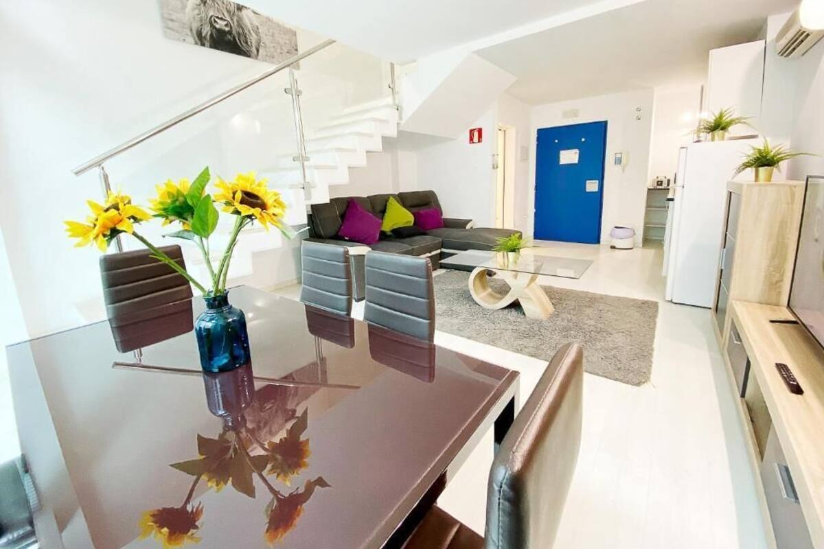 A modern living room with a glass dining table, a sofa with colorful cushions, and a staircase leading to an upper level.