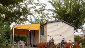 Una cabaña de madera pequeña con un toldo amarillo, dos bicicletas estacionadas afuera y un jardín con flores blancas y púrpuras.