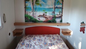 Quarto com cama, cabeceira, prateleira fixada na parede e tapeçaria com tema praiano.
