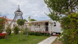 A white mobile home with a balcony, surrounded by a garden with various plants and a decorative metal structure. - Mobile Home 'Casa Gerbera 6' with Private Garden and Wi-Fi (Cavallino-Treporti)