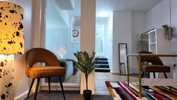 Un salon moderne avec un fauteuil en cuir marron, une plante en pot, un tapis rayé aux couleurs vives et une lampe avec un abat-jour à motifs.