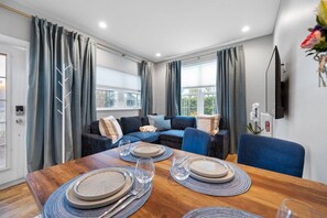 Een moderne woonkamer met een zithoek, een eettafel voor vier personen en een flatscreen televisie aan de muur.
