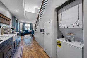 Een compacte keuken met een ingebouwde wasmachine en droger, een zithoek met een blauwe bank, en een trap naar een verdieping erboven.