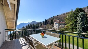 Een balkon met een tafel en stoelen, een fruitschaal en uitzicht op bergen en bomen.