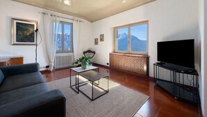 Een woonkamer met een bank, een salontafel, een televisie op een meubel, een radiator, een raam met uitzicht op bergen, en een ingelijst schilderij aan de muur.