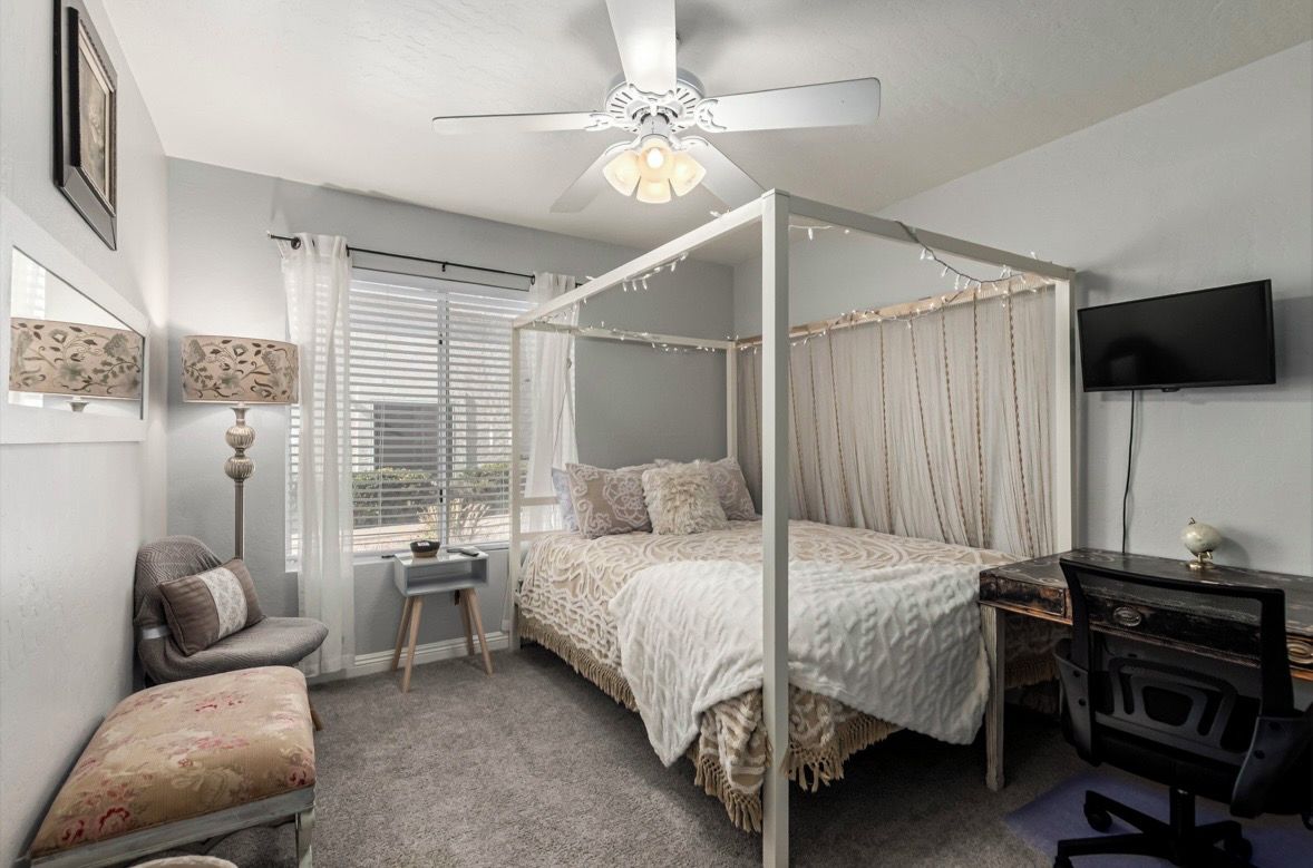 A bedroom with a four-poster bed, a desk with a chair, a television, and a ceiling fan.