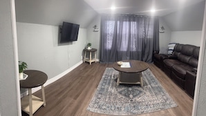 Ein Wohnzimmer mit einem braunen Ledersofa, einem runden Couchtisch, einem an der Wand befestigten Fernseher, einem kleinen Beistelltisch mit einer Pflanze und einem gemusterten Teppich.