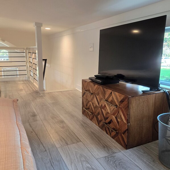 A modern living room with a flat-screen TV on a wooden cabinet, a sofa, and a window with a view of the outdoors.